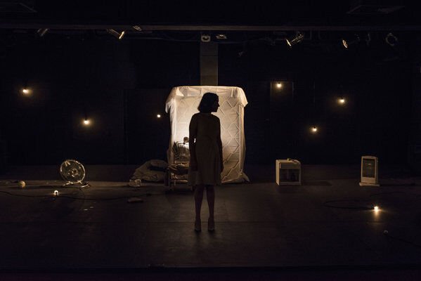 Escena en penumbras, con algunas lámparas que apenas se ven encendidas en sobre el fondo. En el centro un colchón parado, apoyado sobre la pared del fondo, iluminado más que el resto de la escena, en el frente, se ve la silueta en sombras de una mujer de pie mirando hacia un lado.