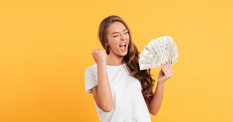Joven mujer pelirroja, con la boca abierta como gritando y con actitud triunfalista mirando un abanico de billetes y celebrando, remera blanca sobre fondo amarillo.